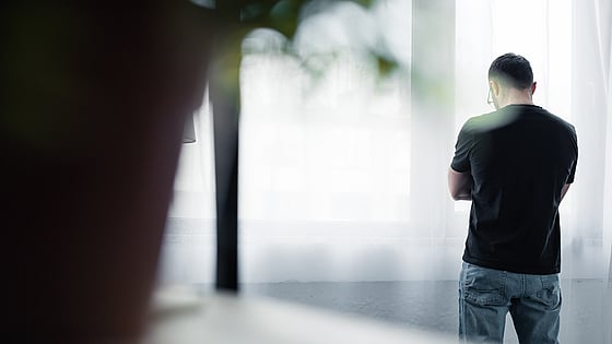 Bild tagen bakifrån på en man som står med armarna i kors vid ett fönster. Foto: Mostphotos